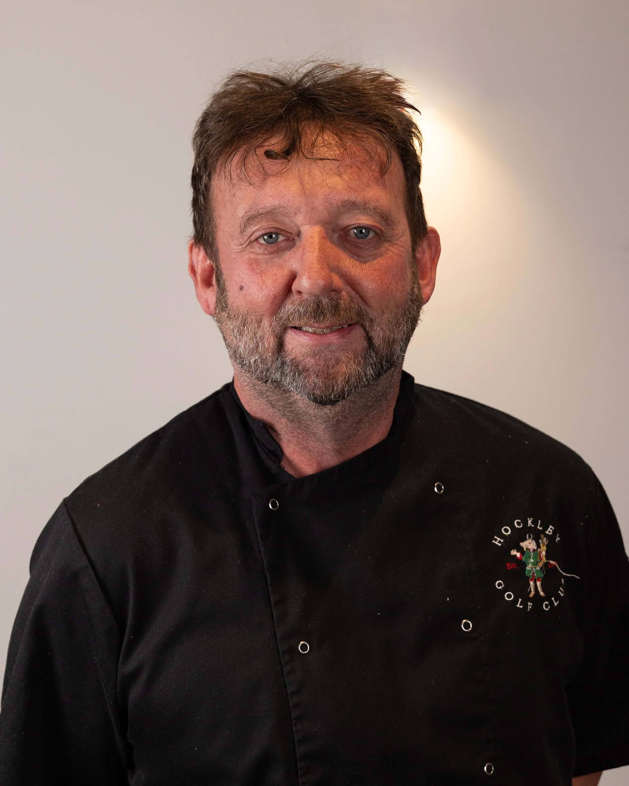 A middle-aged man with a beard and disheveled hair wearing a black chef's coat with an embroidered logo of a golf club on the chest, standing against a plain wall.
