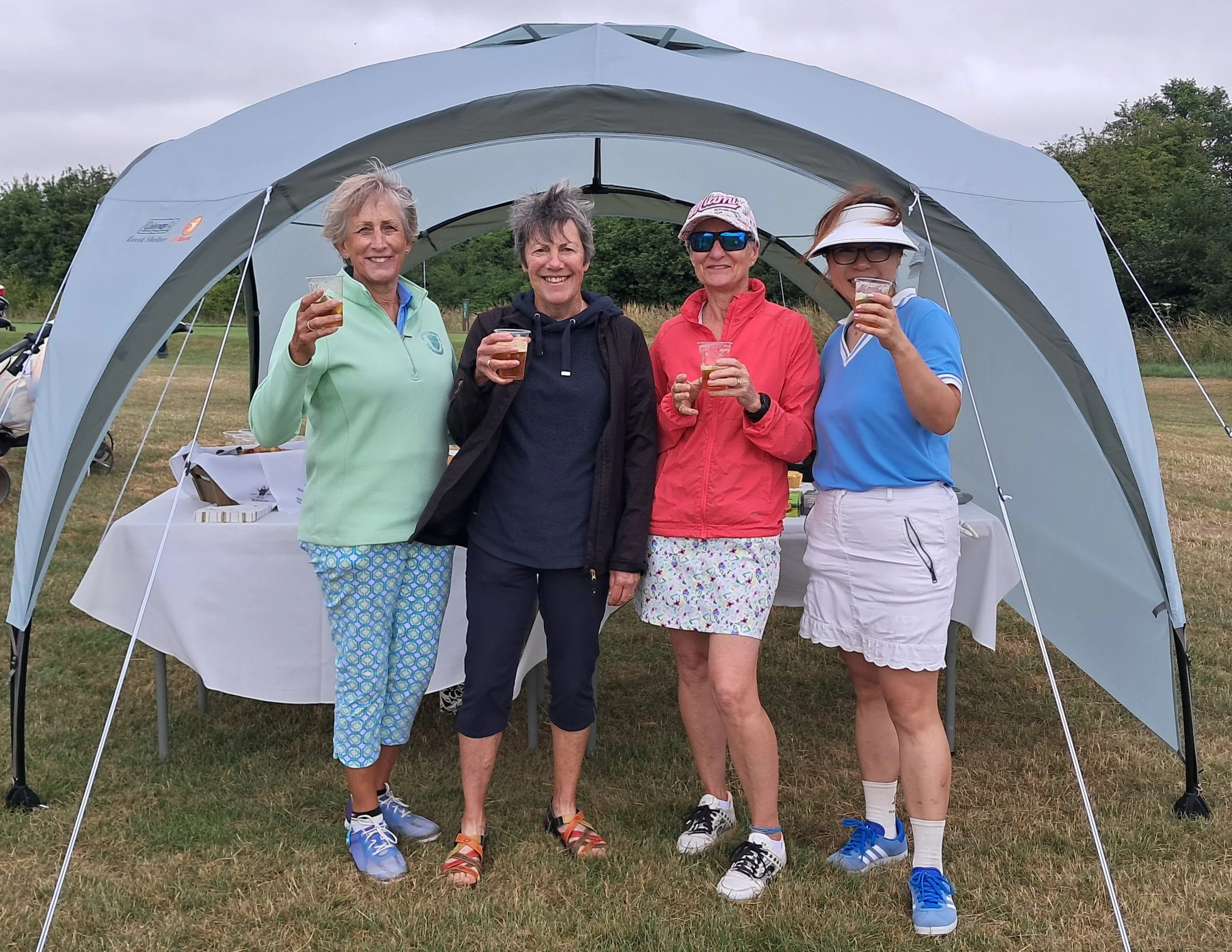 Four women standing in front of a camping shelter, holding drinks and smiling. They are outdoors on a grassy area with trees in the background. They appear to be enjoying a social gathering or outdoor event.
