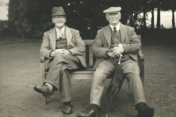 Two older men sitting on a park bench outdoors, dressed in vintage clothing including hats and suits.