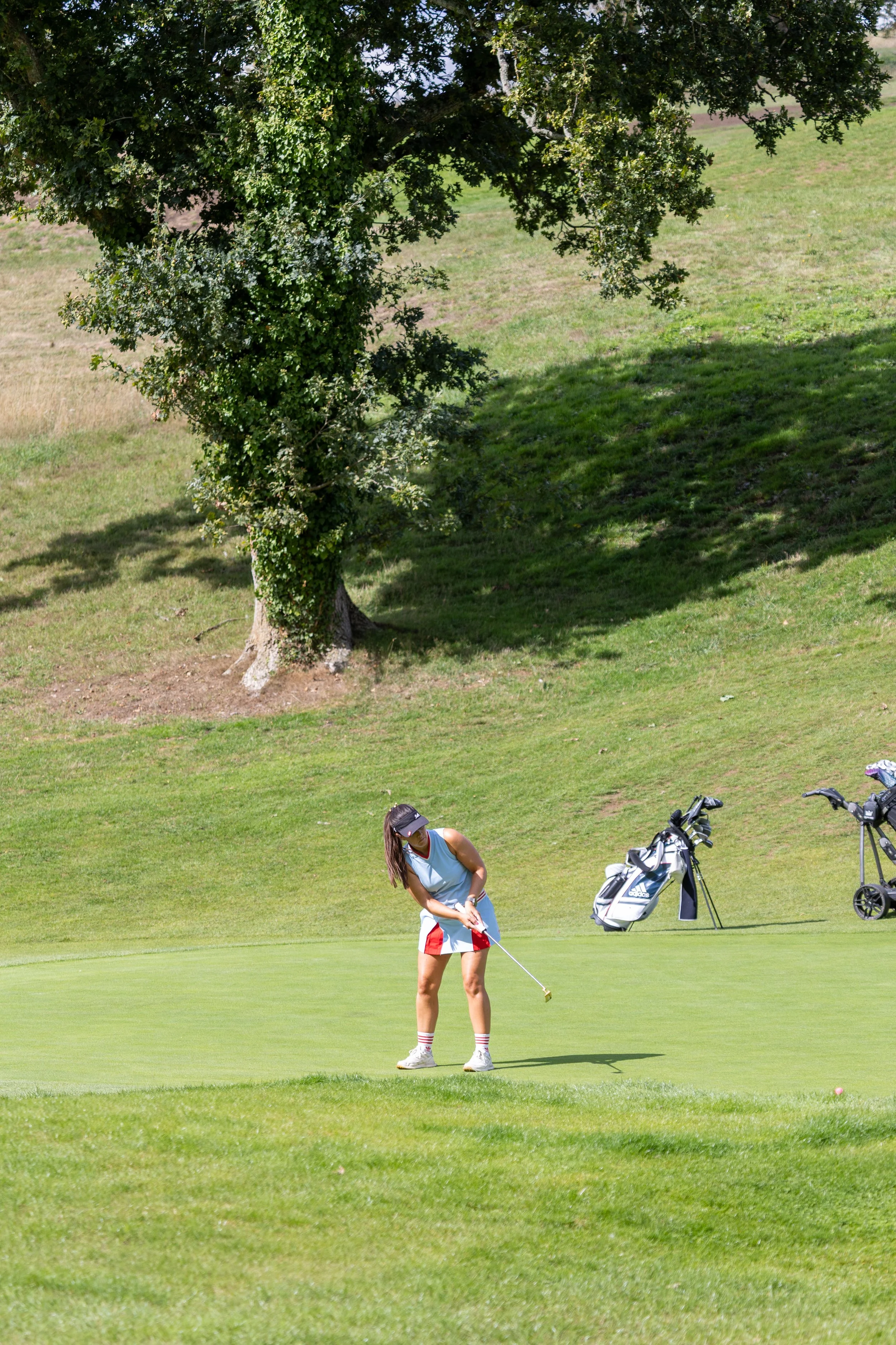 A woman playing golf on a lush green course, identifying her golf clubs nearby, with a large tree providing shade in the background.
