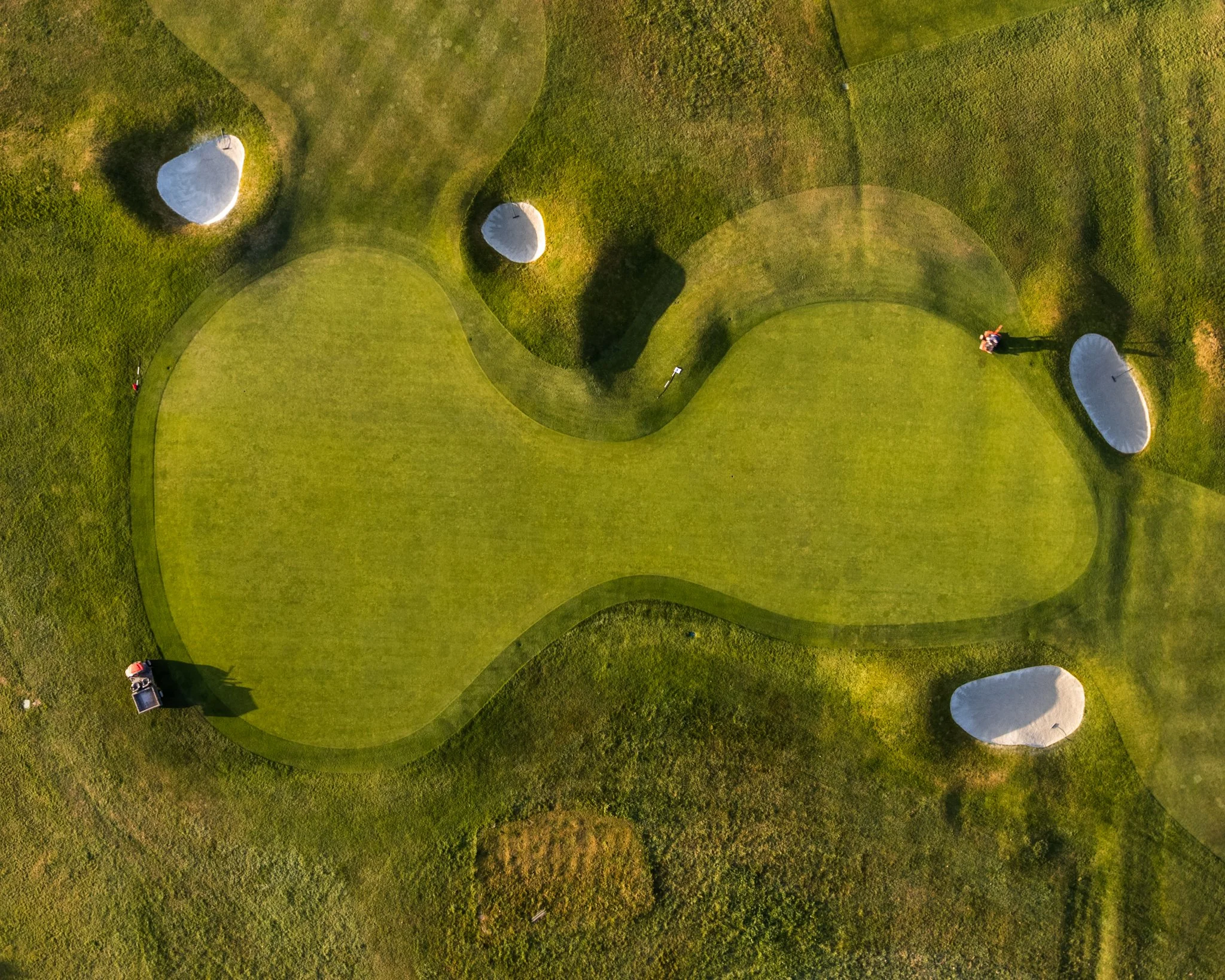 Aerial view of a golf course with a green surrounded by sand bunkers and lush grass, showing two golfers on the course and a golf cart.
