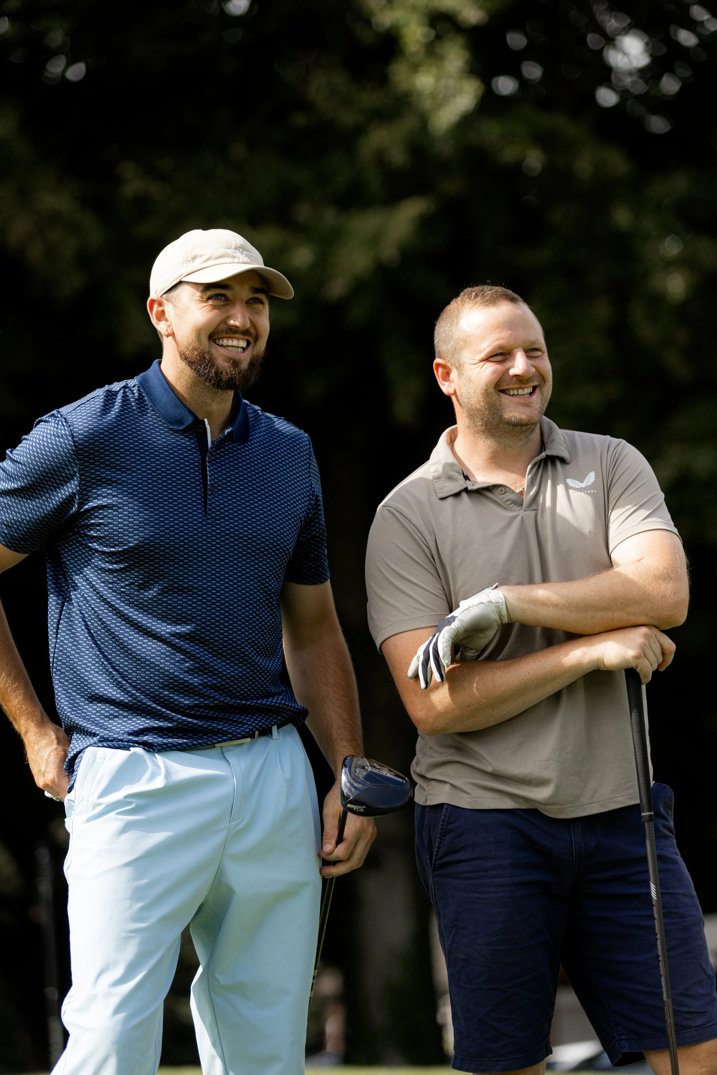 Two men standing outdoors on a golf course, smiling and enjoying the day, with golf clubs in hand.