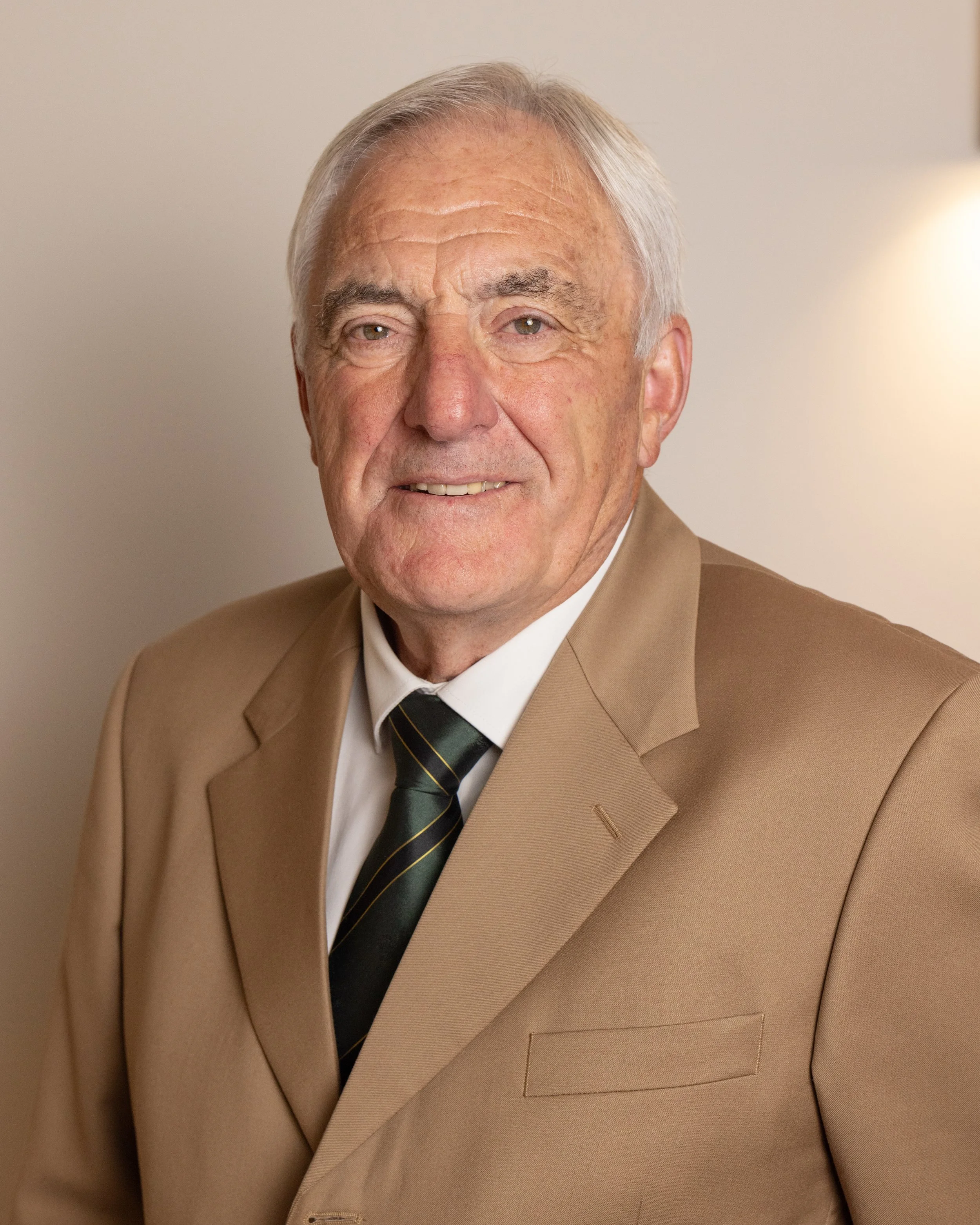 Portrait of an elderly man with gray hair wearing a beige suit, white shirt, and black tie, standing against a plain wall.