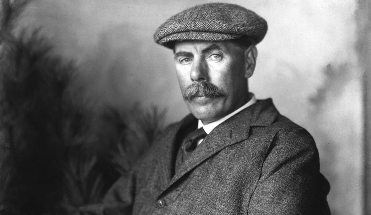Black and white portrait of a man with a mustache wearing a tweed cap and a suit with a tie, looking at the camera.