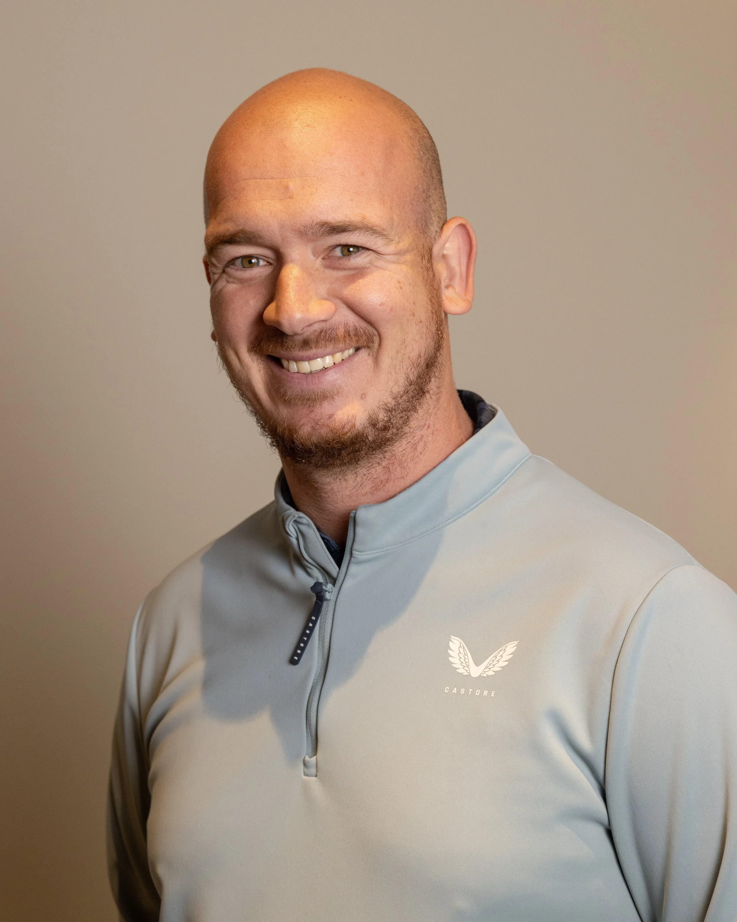 A smiling man in a light gray sports jacket with a logo on the chest, standing against a neutral background.