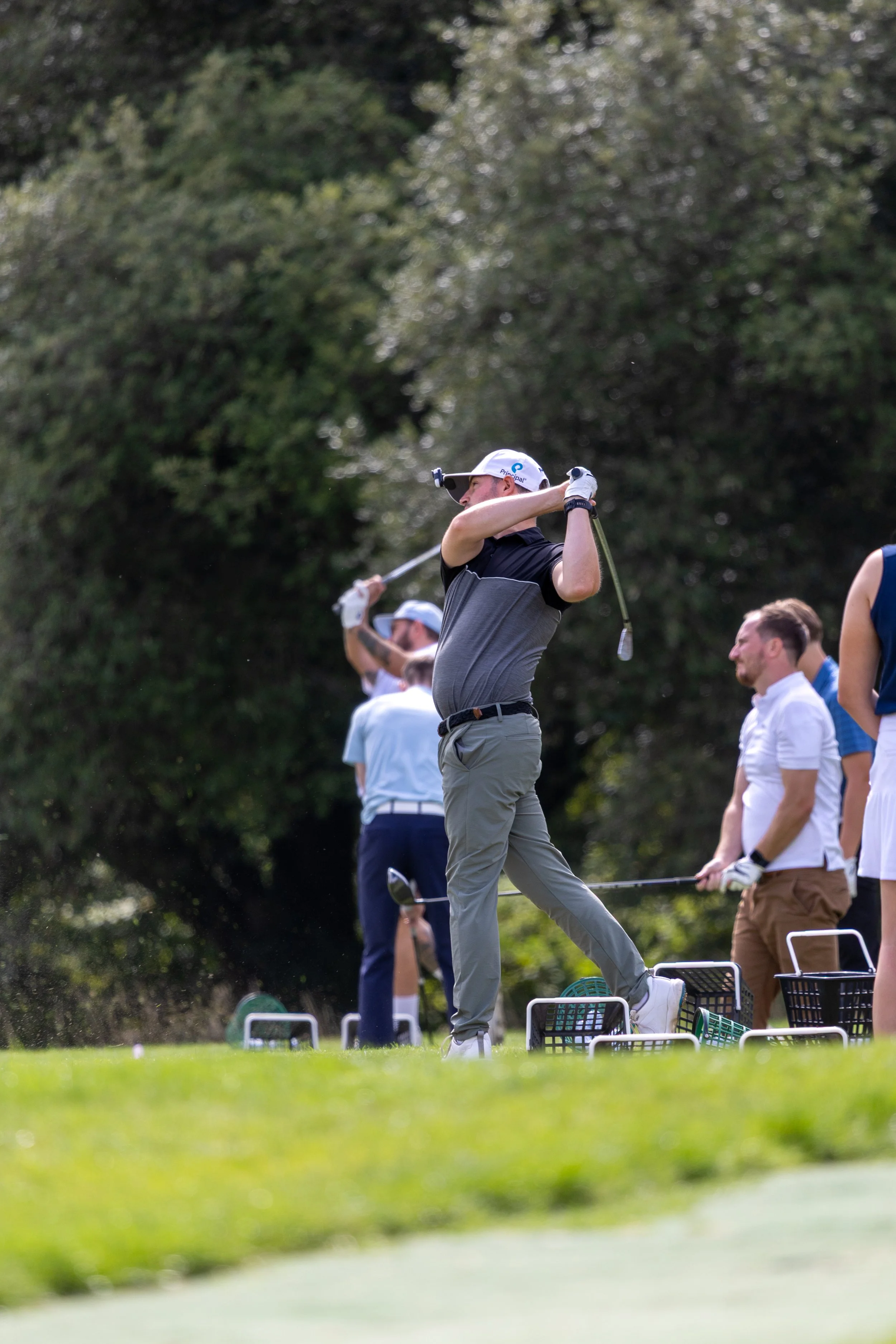 Golfers on a golf course, with one man swinging a golf club in the foreground and others watching or preparing in the background.