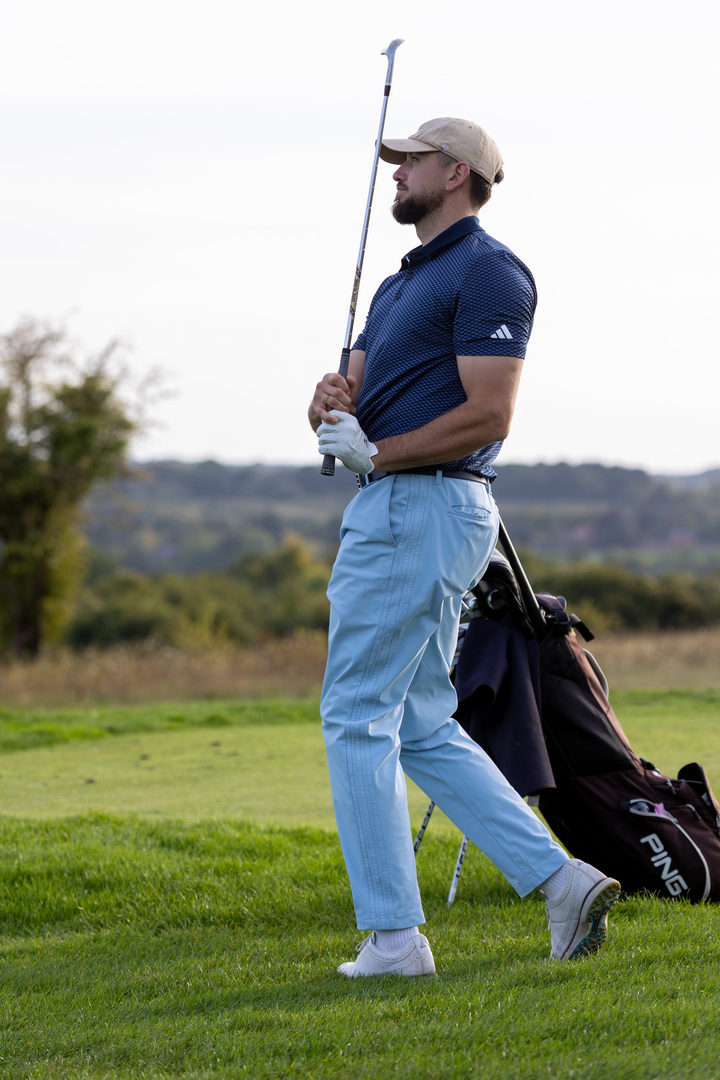A man standing on a golf course, holding a golf club, with a golf bag behind him.
