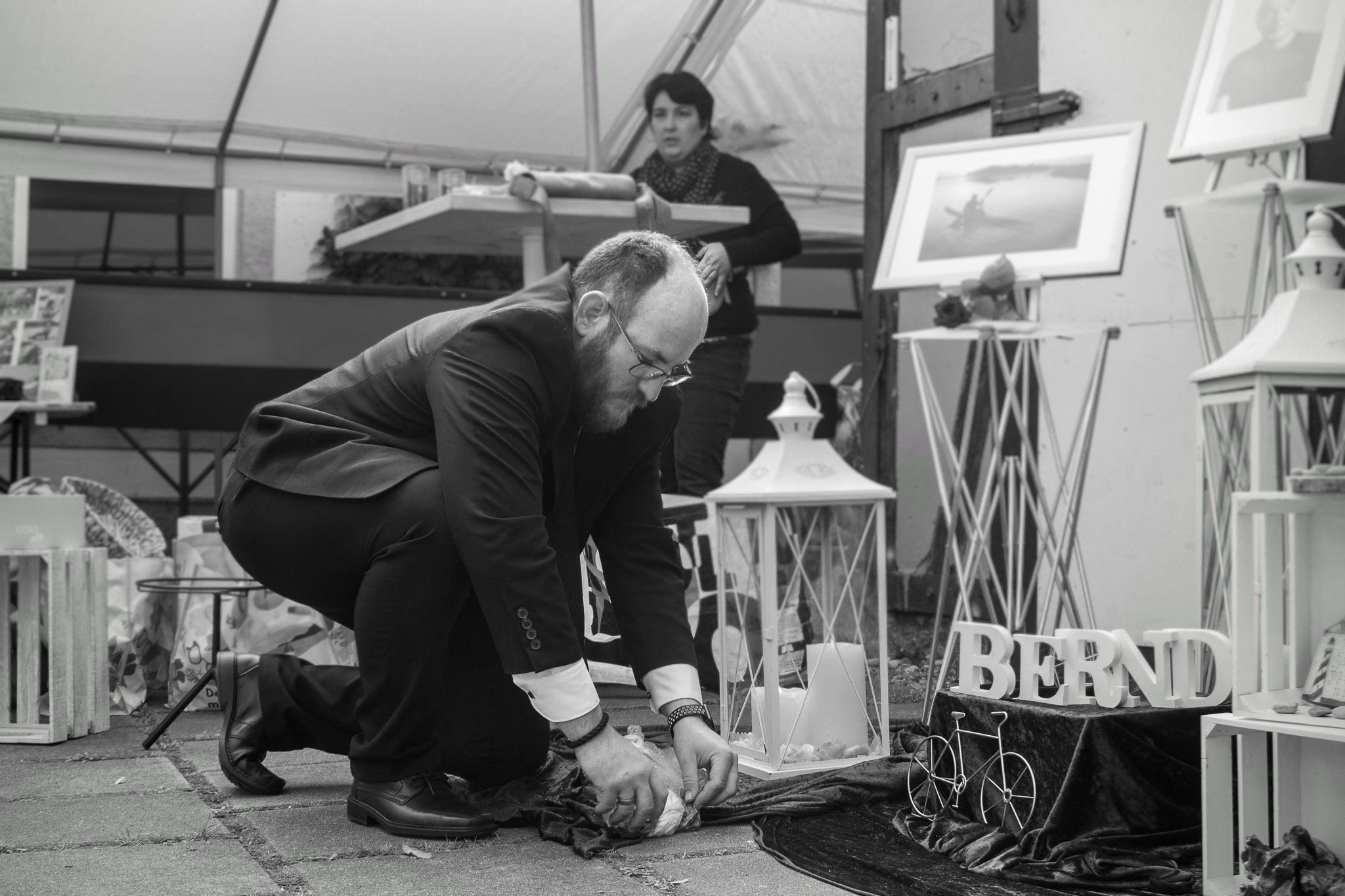 Ein Mann auf Knien beim Dekorieren eines Standes, im Hintergrund eine Frau, umgeben von Bildern und Dekorationsgegenständen, in einer Kunst- oder Flohmarkt-Umgebung.