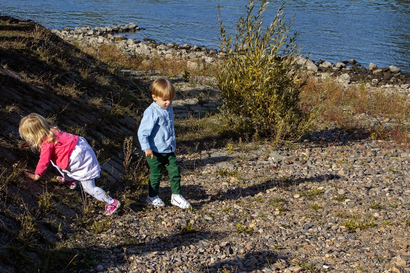 Zwei Kinder, ein Mädchen in rosa und weiß, ein Junge in blau und grün, spielen am Ufer eines Sees mit Steinen und Pflanzen.