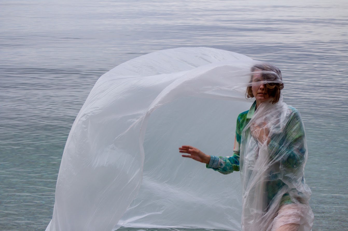 Eine Frau trägt einen transparenten Kunststoff-Überzug an einem ruhigen See, der Wind bläst den Überzug leicht um sie herum.