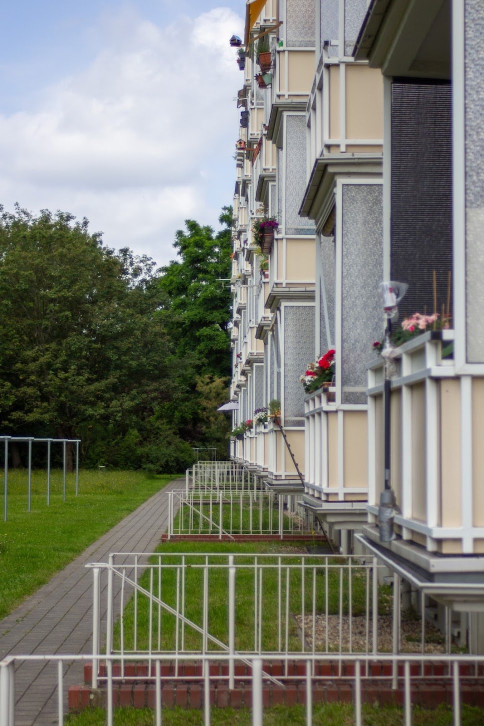 Mehrstöckiges Wohngebäude mit blumengeschmückten Balkonen, neben einer grünen Wiese und Bäumen, bei bewölktem Himmel.