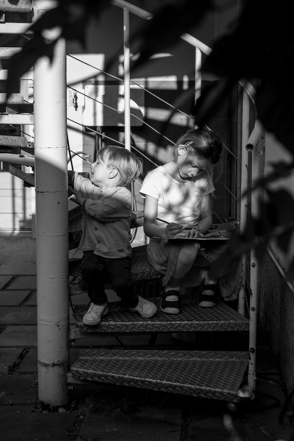 Schwarz-Weiß-Foto von zwei Kindern, einem Jungen und einem Mädchen, auf einer Metalltreppe draußen, das Mädchen schreibt oder zeichnet in ein Notizbuch, während der Junge sich an einer Stange festhält, um die Treppe hinaufzusteigen.