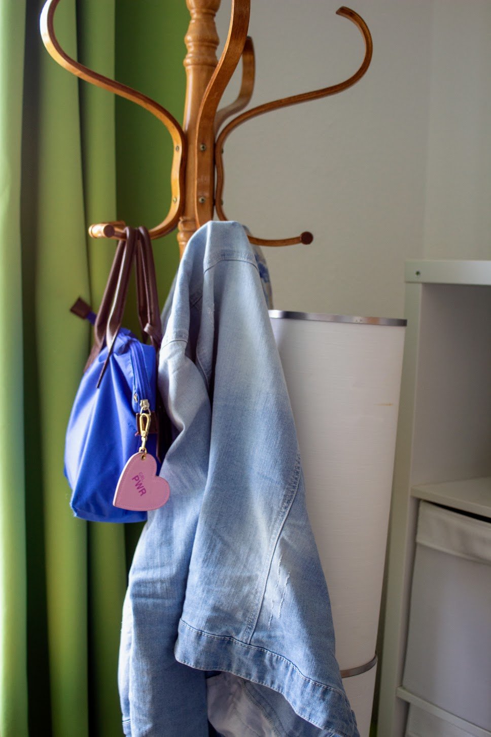 Eine Garderobe mit drei Haken, auf denen eine blaue Tasche, eine Jeansjacke und ein Regenschirm hängen. Im Hintergrund grüne Gardinen.