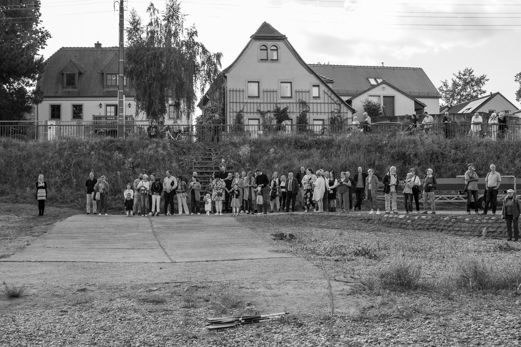 Eine Gruppe von Menschen, die in einer Warteschlange an einem Uferbereich stehen, im Hintergrund Häuser und Bäume, schwarz-weiß Foto.