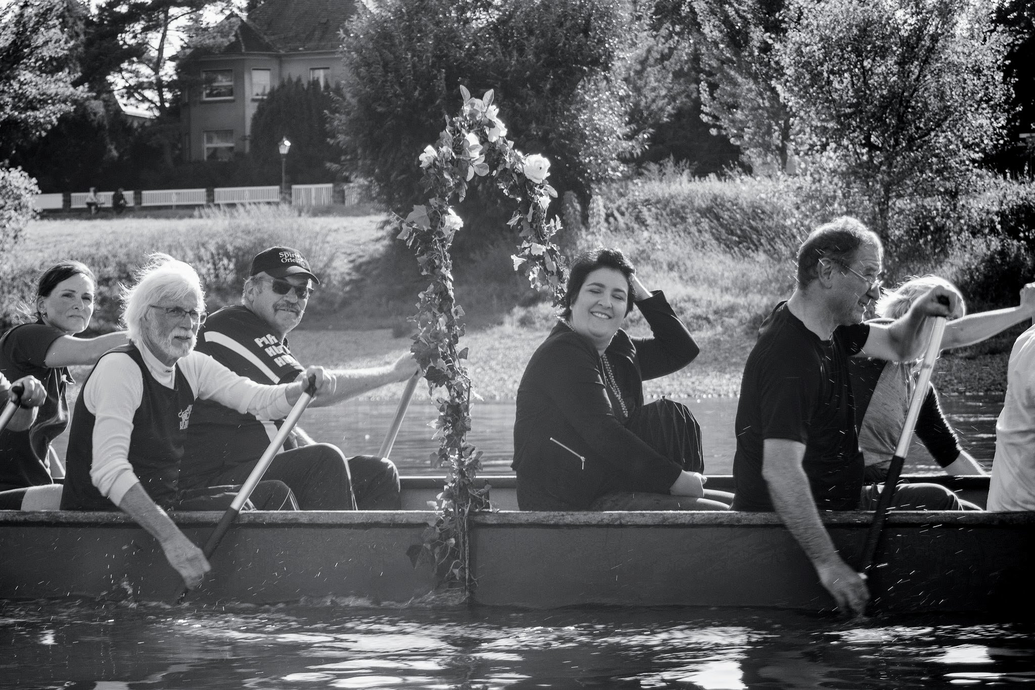 Menschen paddeln mit einem Boot auf einem Fluss, in der Mitte lächelt eine junge Frau, im Hintergrund Bäume und Häuser, Schwarz-Weiß-Fotografie.