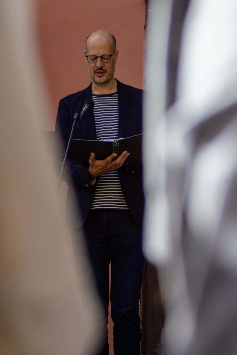 Mann liest vor einem Mikrofon, umgeben von mehreren Personen, hinter einer roten Wand.