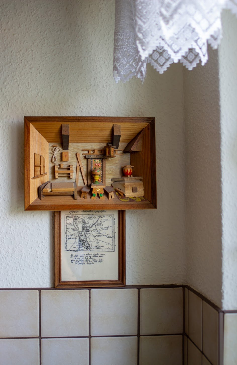Holzspielzeug in einem Wandregal, das ein kleines Haus oder eine Krippe darstellt, mit einem Bild darunter an einer weißen Wand, teilweise hinter Fliesen.