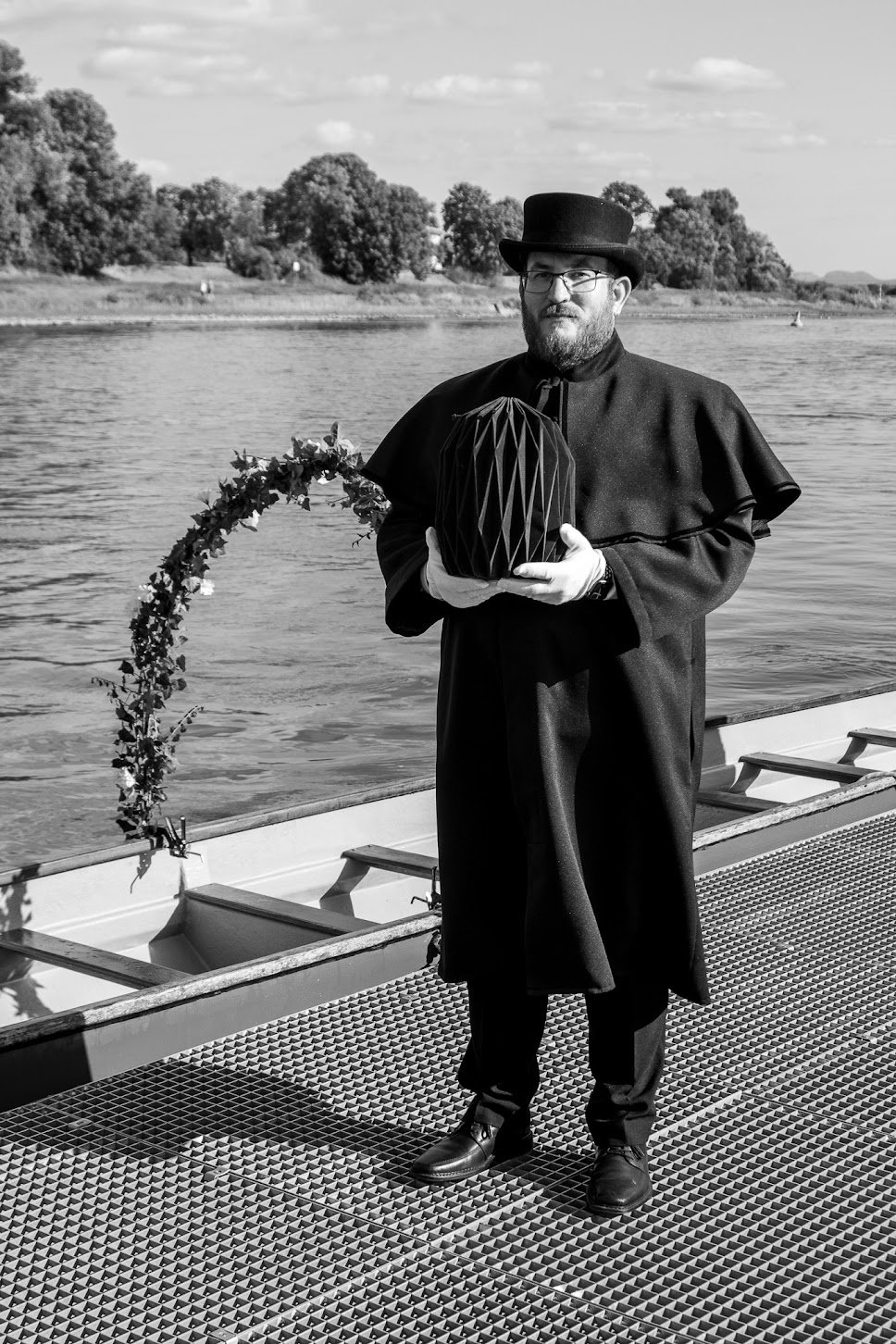 Mann in historischer Kleidung mit Zylinder und Umhang steht auf einem Steg am Fluss, hält eine dekorative Laterne, im Hintergrund Bäume und Wasser.
