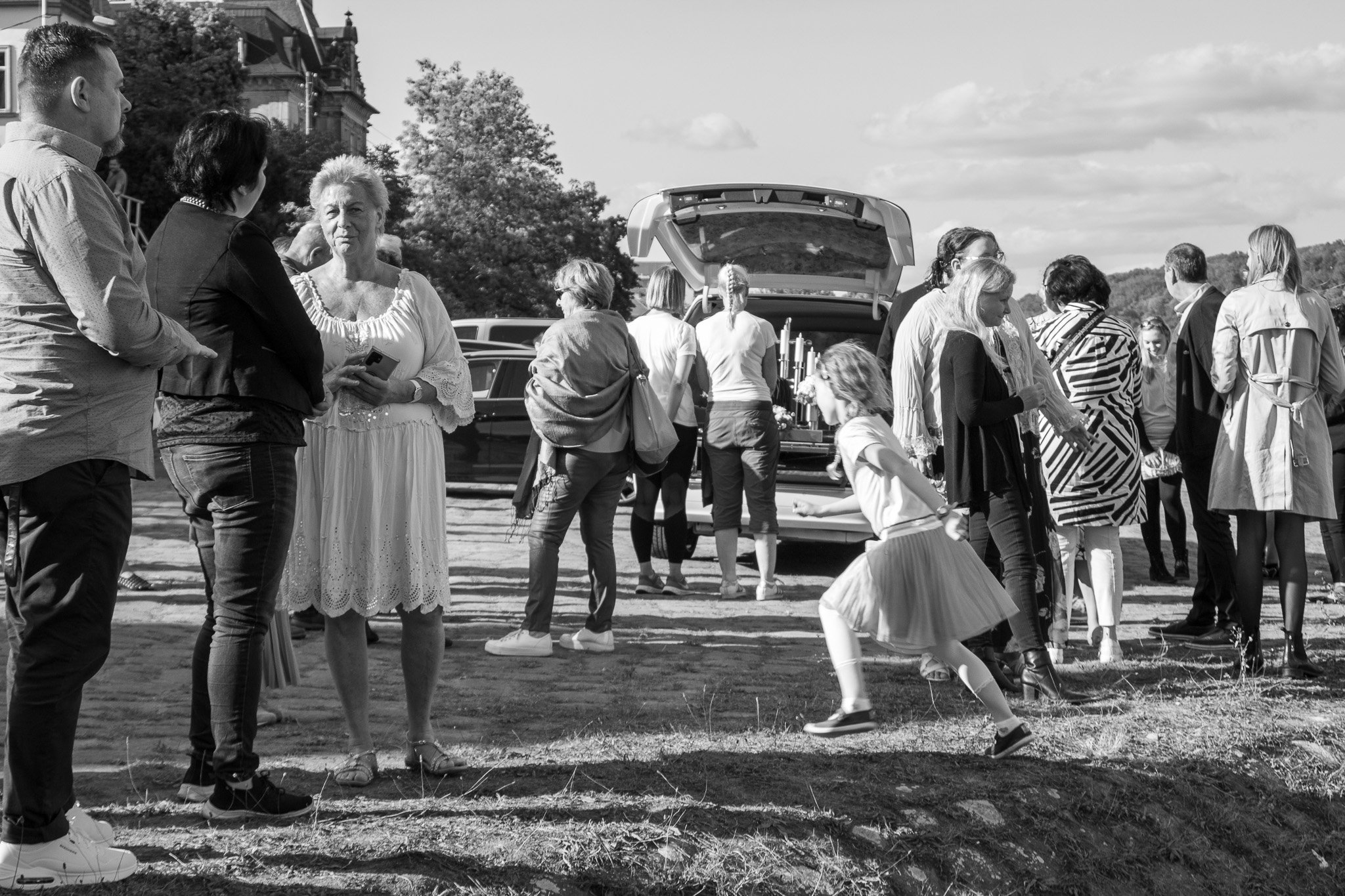 Mehrere Menschen versammeln sich im Freien, einige stehen in Gruppen, während eine junge Frau in Sommerkleid feiert und einen Sprung macht. Im Hintergrund sind Autos und Bäume sichtbar.