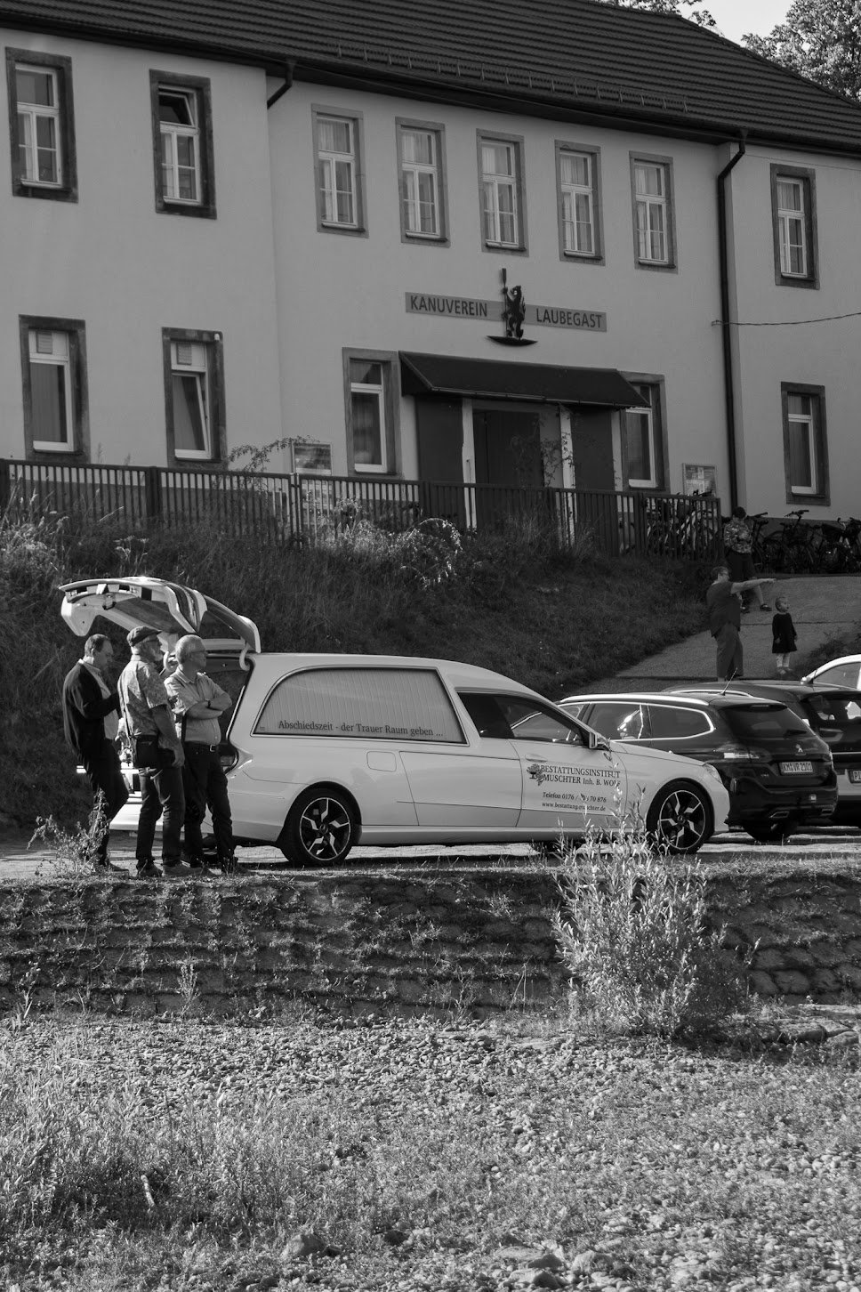Ein Schwarz-Weiß-Foto zeigt eine kleine Gruppe von Menschen um ein Fahrzeug mit Sarg auf einem Parkplatz vor einem Gebäude mit dem Schild 'Kanuverein Laubegast'. Im Hintergrund sind Kinder und Erwachsene zu sehen, einige mit Fahrrädern.