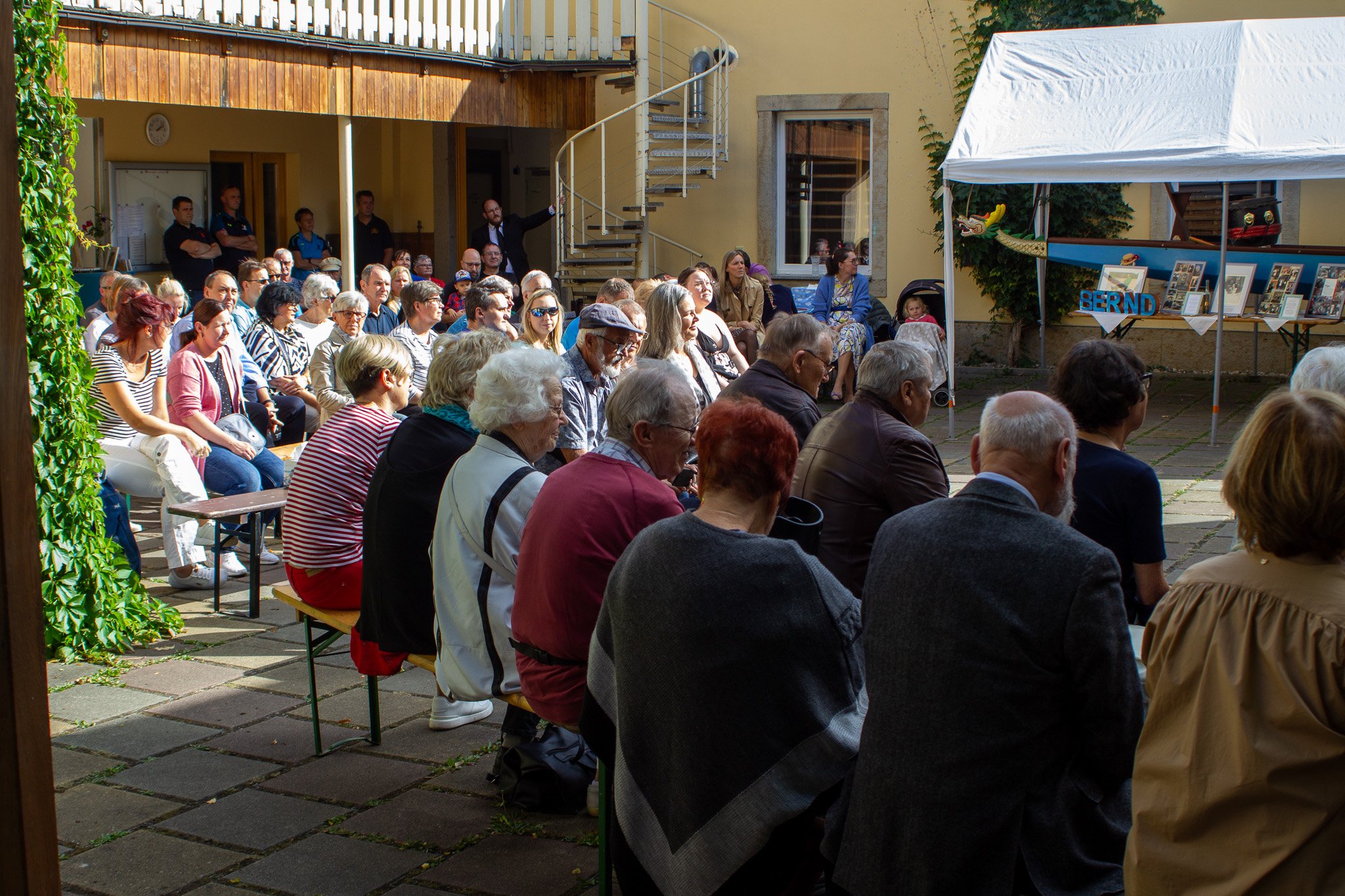 Menschen sitzen im Schatten bei einer Trauerfeier, einige stehen im Hintergrund, ein weißer Pavillon mit Fotos und Buchstaben, die den Namen 'Bernd' zeigen, im Hintergrund.