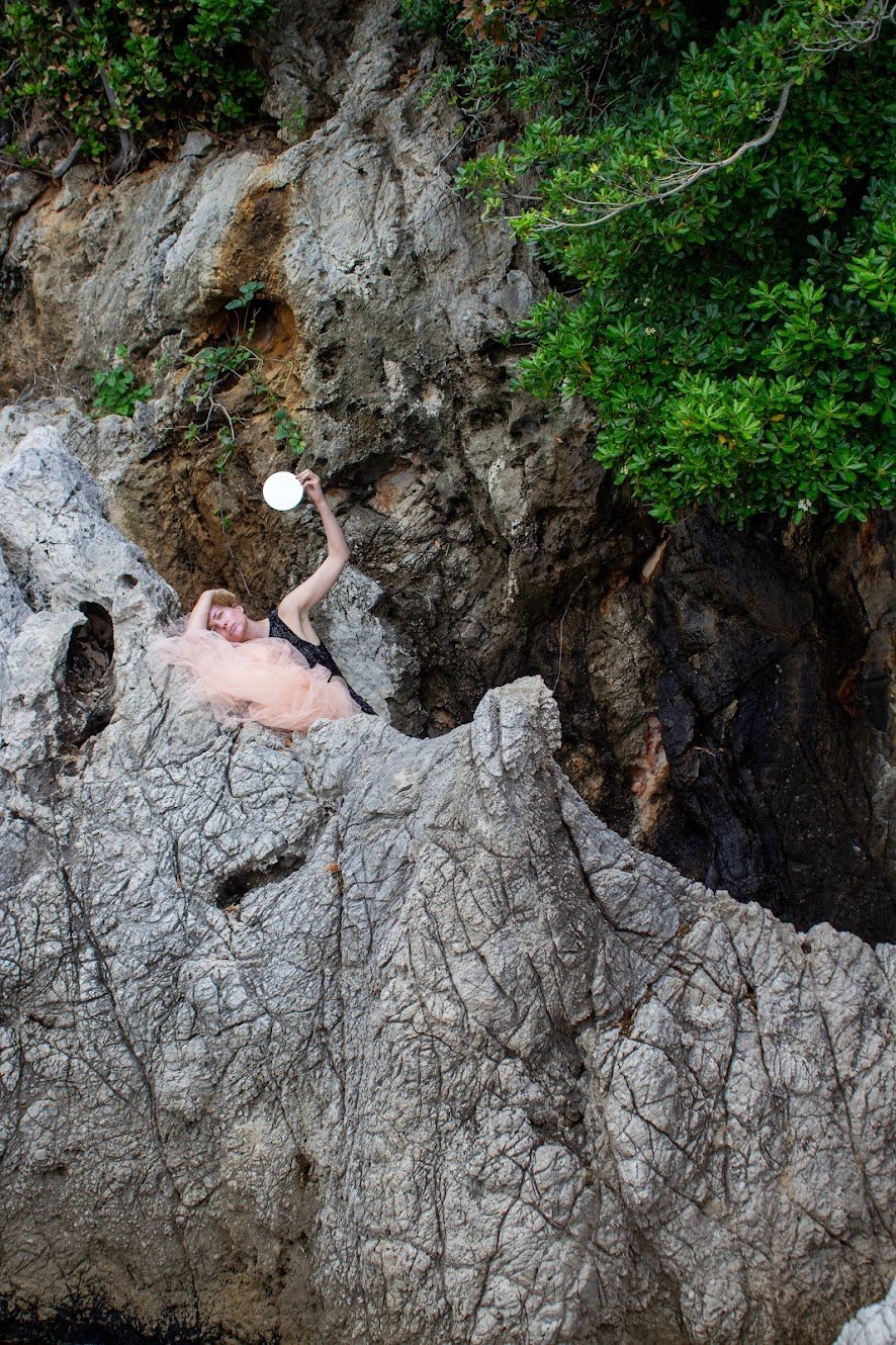 Frau in einem Tüllkleid liegt auf großen Felsen vor einer felsigen Klippe, umgeben von grünen Büschen, und hält einen Spiegel in die Luft.
