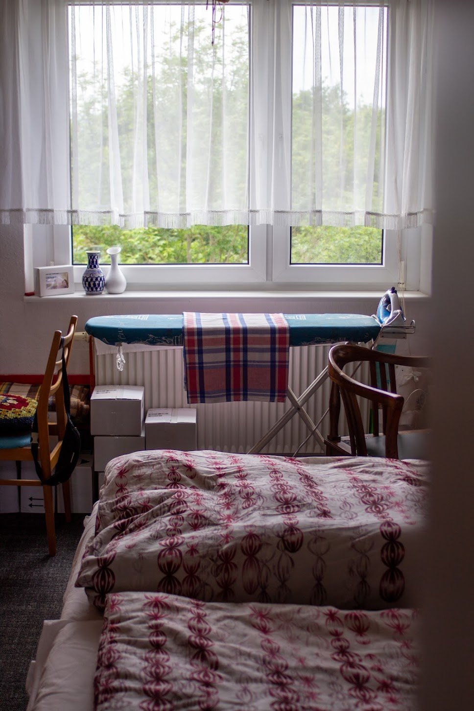 Ein Schlafzimmer mit einem Fenster, einem Bügelbrett mit einem karierten Tuch, einem Bett mit blumigem Bettzeug, einem Stuhl, einem Nachttisch und Dekorationen auf der Fensterbank.