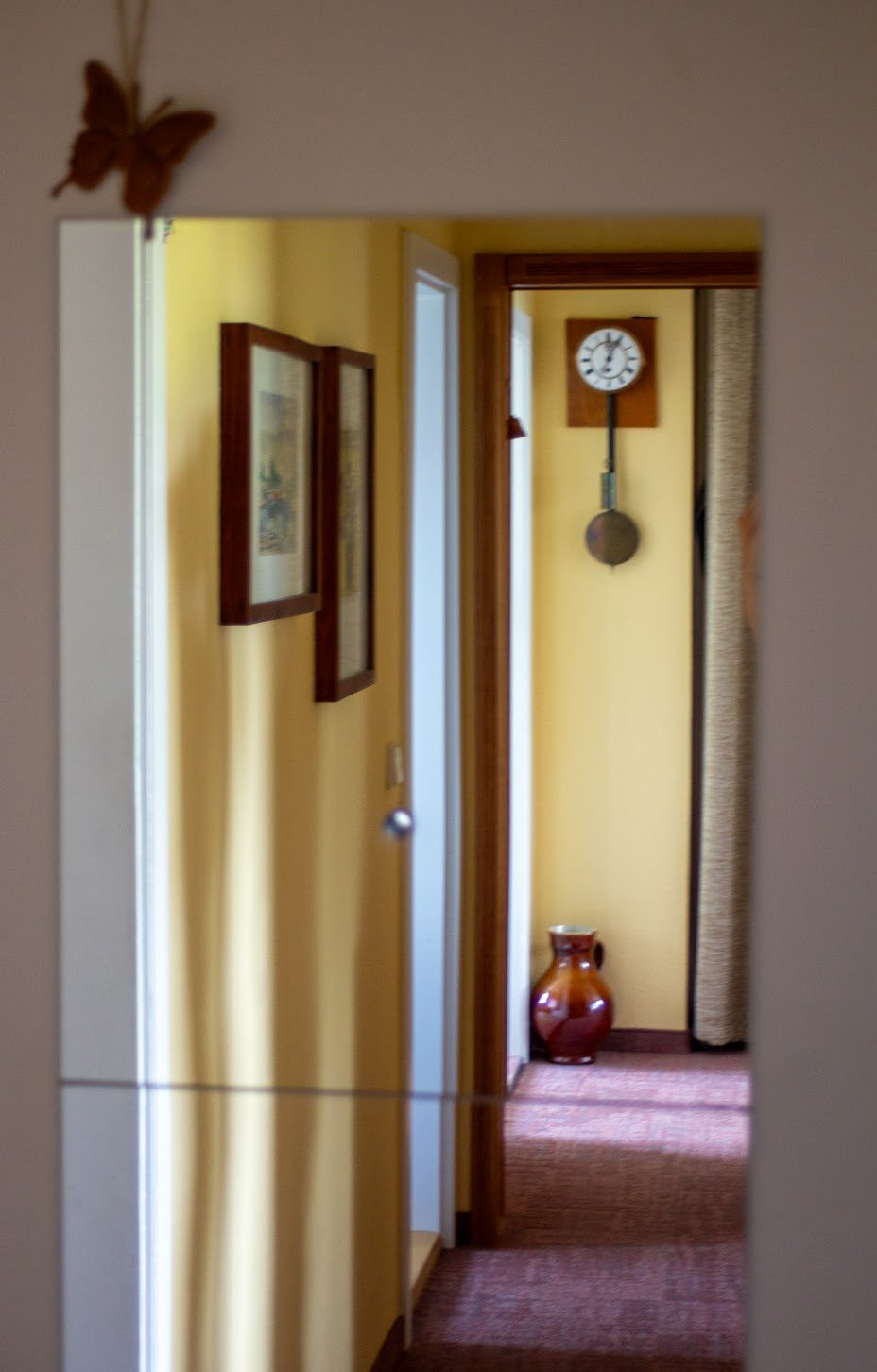 Blick durch einen Spiegel in einen Flur mit gelben Wänden, zwei Bilder an der Seite, ein Wandklavier, eine antike Vase auf dem Boden, eine Uhr am Ende des Flures mit Pendel.
