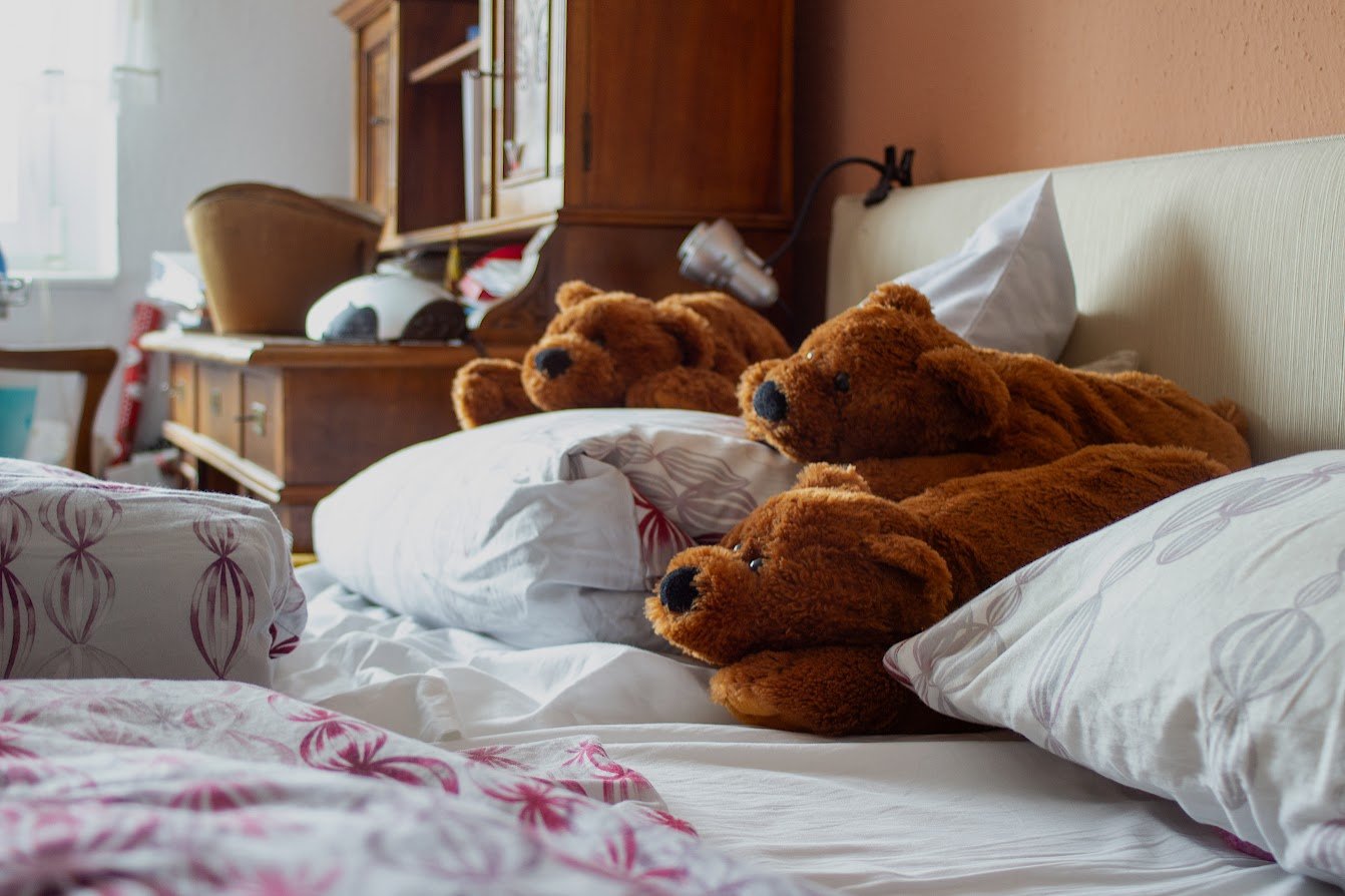 Bett mit vier braunen Teddybären und Kissen im Schlafzimmer, Holzregal im Hintergrund.