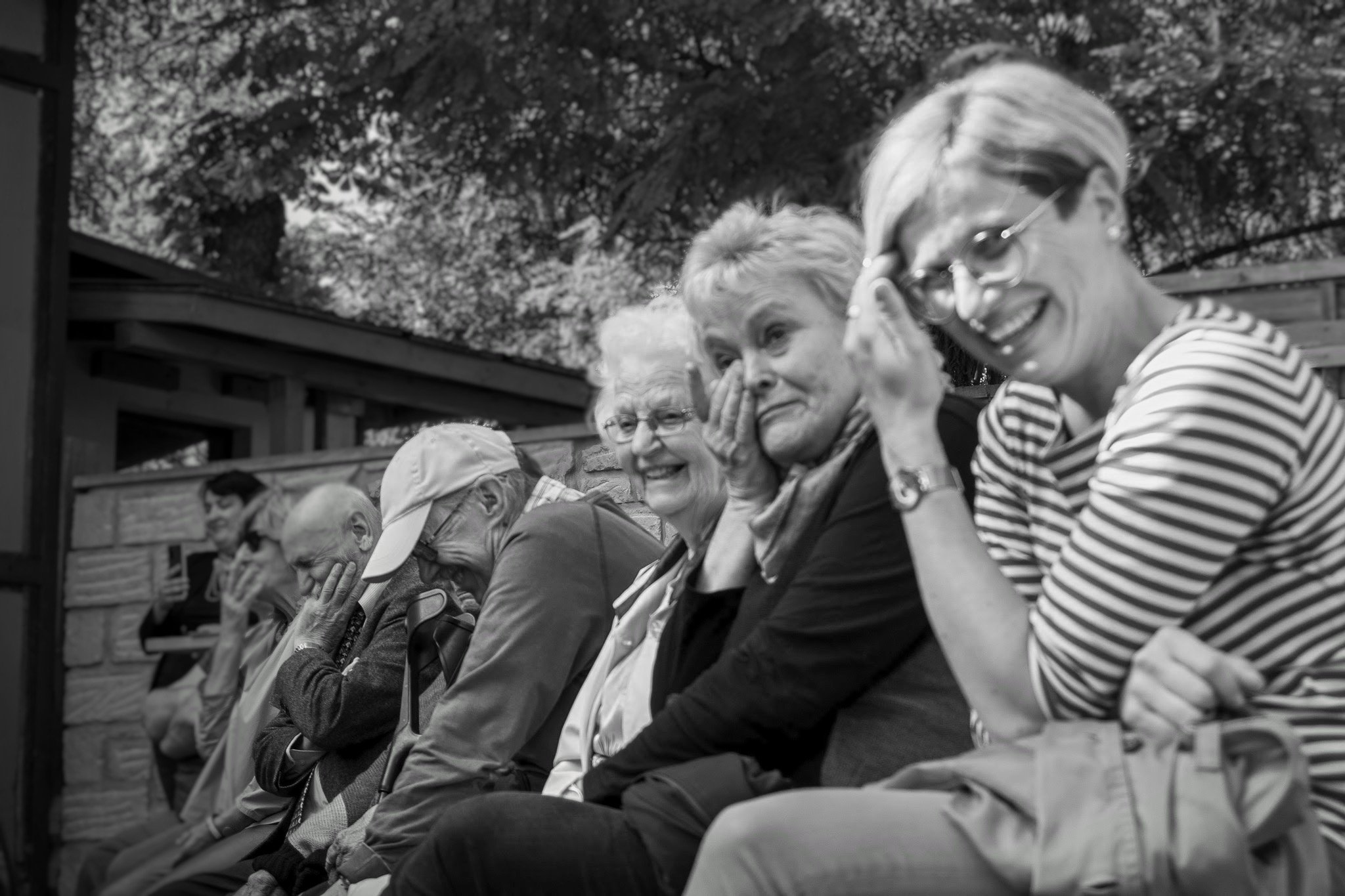 Eine Gruppe älterer Menschen sitzt zusammen im Freien und lacht, einige lachen herzlich, während andere ihre Augen vor Freude schließen.