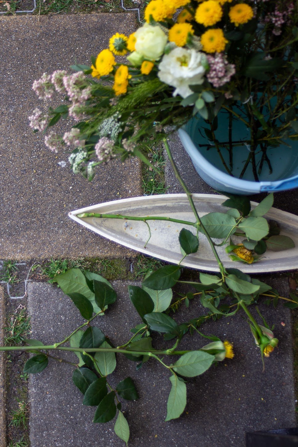 Blumen, darunter gelbe, weiße und rosa, auf einer Gehwegplatte, einige liegen in einer weißen Vase.