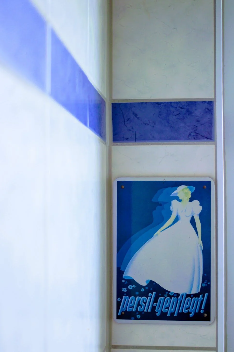 Ein Wandbild mit einer Frau in einem weißen Kleid und einem Hut, das gedreht wirkt, in blau-weißer Farbgestaltung, mit dem deutschen Text "persil-geleitet!" darunter.
