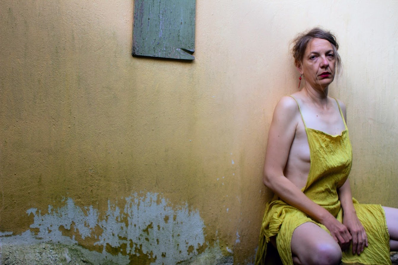 Frau sitzt mit zerrissenem gelbem Kleid in einem Raum mit gelber Wand und bunter Wandkunst.