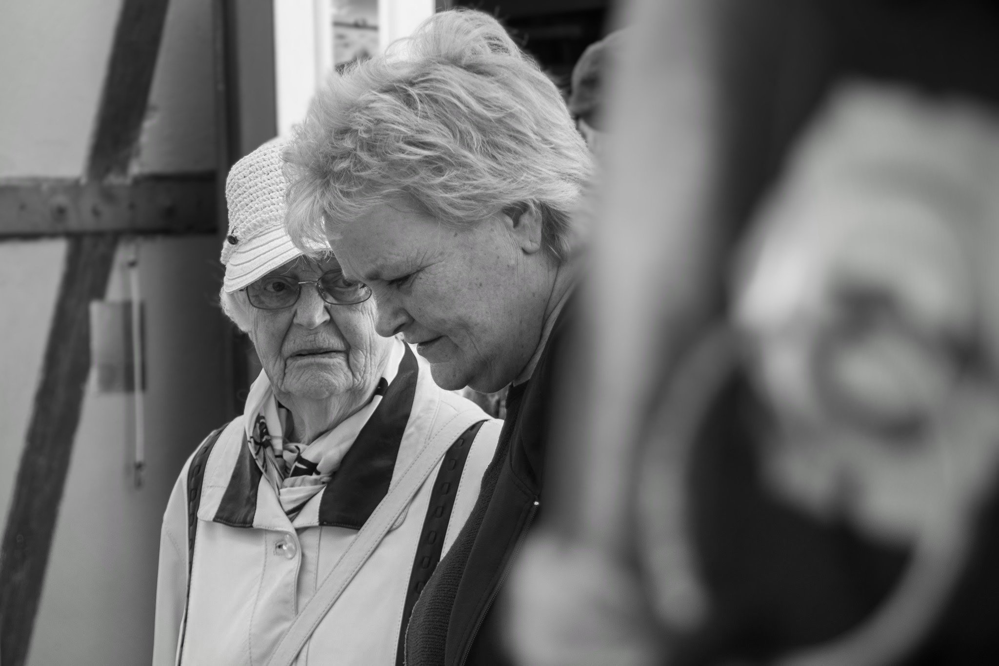 Zwei ältere Frauen mit kurzen, grauen Haaren, eine trägt eine Sonnenbrille und einen Sonnenhut, die andere hat einen schwarzen Schal und eine weiße Jacke, stehen im Gespräch in einem Raum mit Holzrahmen an der Wand.