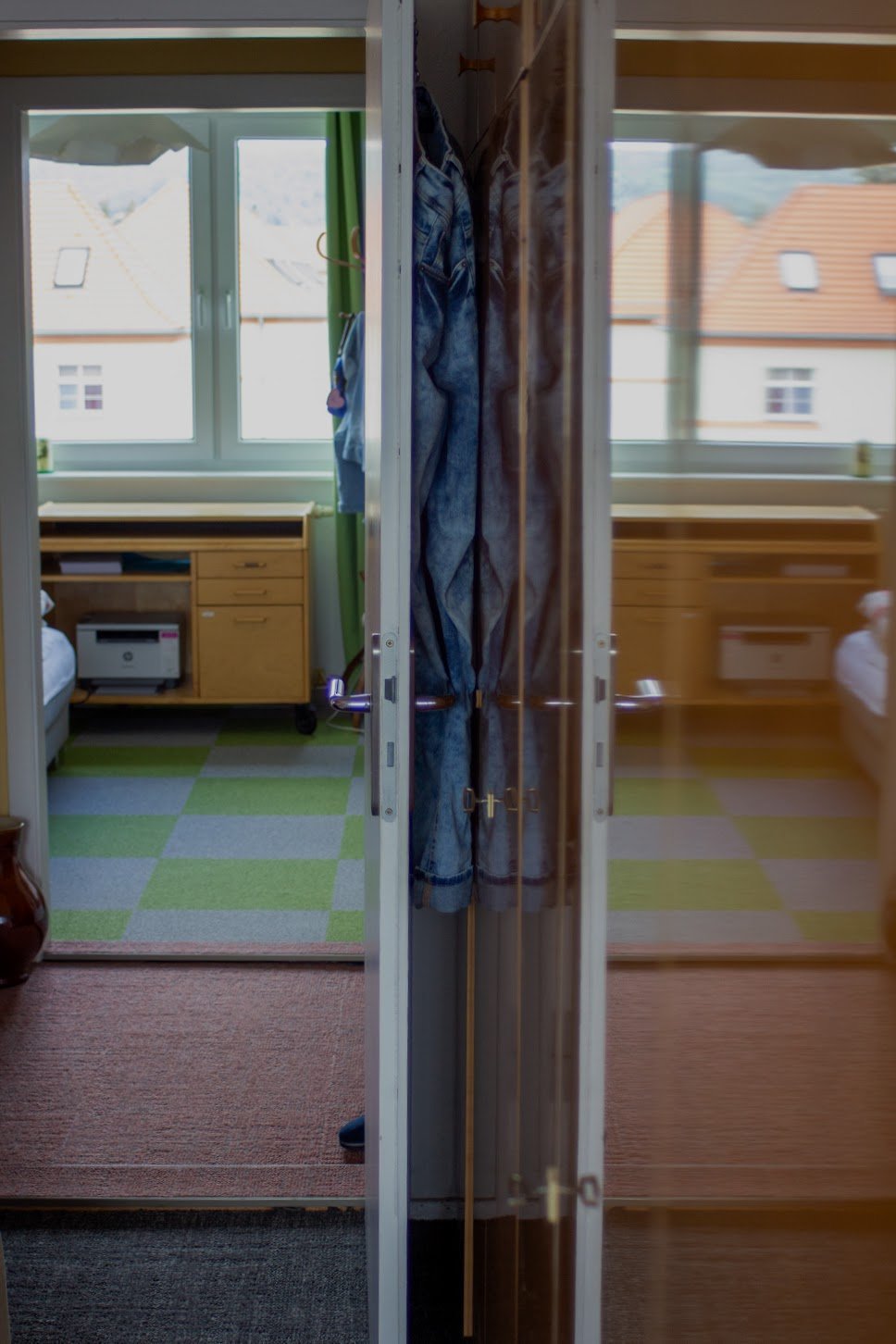 Blick in ein Zimmer, das durch eine offene Schiebetür mit Spiegelhülle getrennt ist. Im Hintergrund sind Fenster mit Blick auf Häuserdächer, eine Kommode mit Computer sowie Kleidung an der Tür zu sehen.
