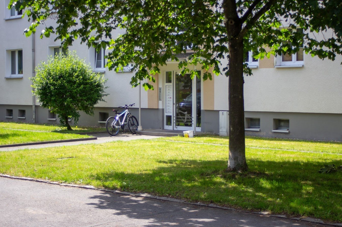 Mehrfamilienhaus mit Fahrrad vor Eingang, umgeben von grünen Bäumen und Rasen