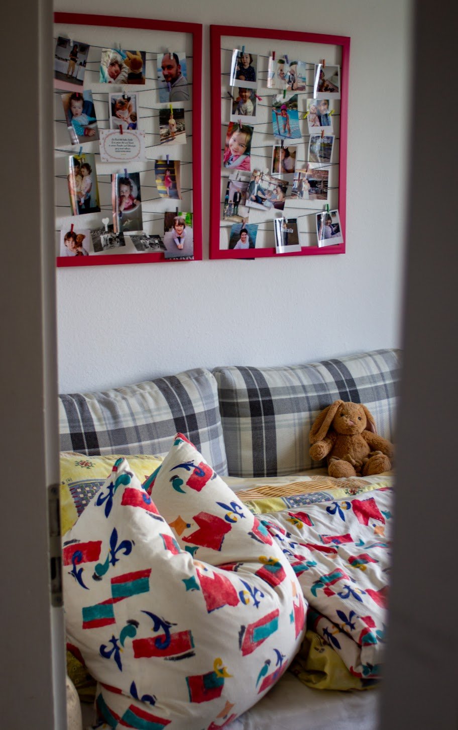 Blick durch eine Tür in ein Wohnzimmer mit einem gepolsterten Sofa, bunten Kissen, einem Plüschhasen und einer Fotowand mit Familienbildern.