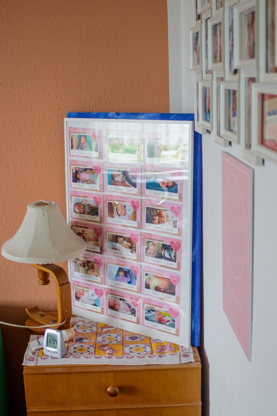 Eine Tafel mit Fotos von Babys, dekoriert mit pinken Herzen, steht neben einem Nachttisch mit einer Stecklampe und einem digitalen Thermometer. An der weißen Wand sind mehrere kleine Bilderrahmen angebracht.