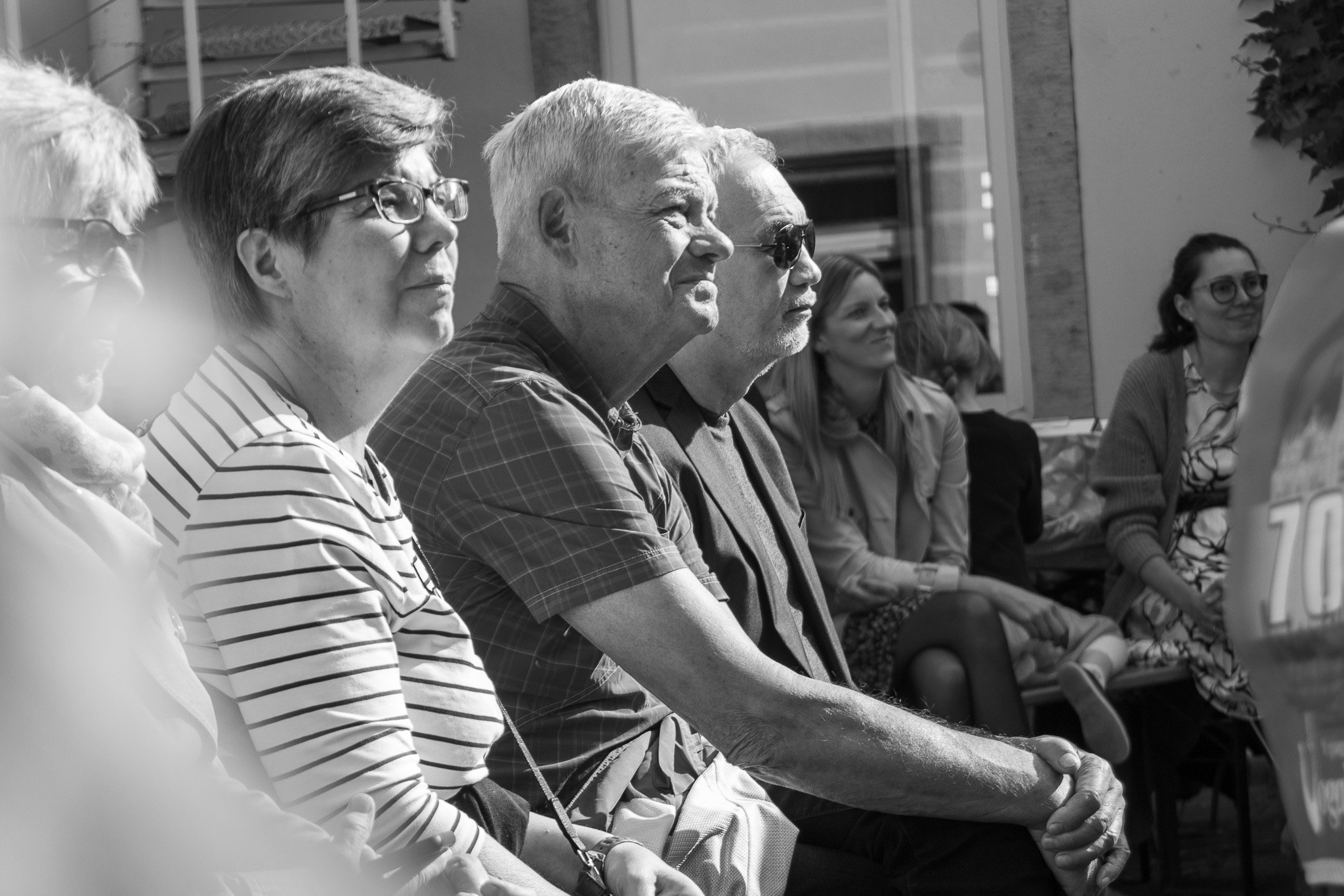 Mehrere Menschen sitzen in einer Reihe bei einer Veranstaltung im Freien, schwarz-weiß Fotografie.