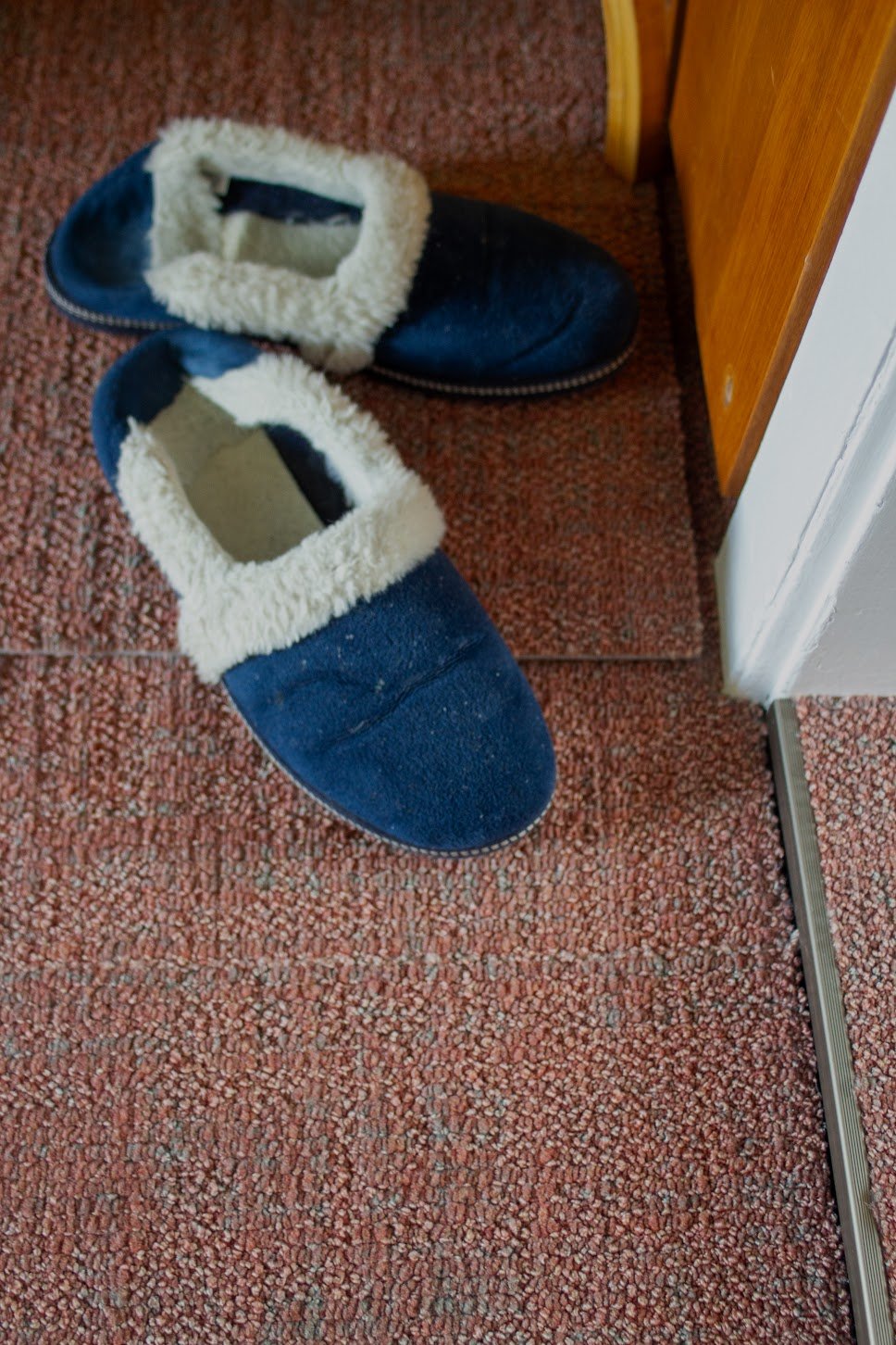 Ein Paar blaue Hausschuhe mit weißem, flauschigem Innenfutter auf einem roten Teppichboden neben einer Tür.