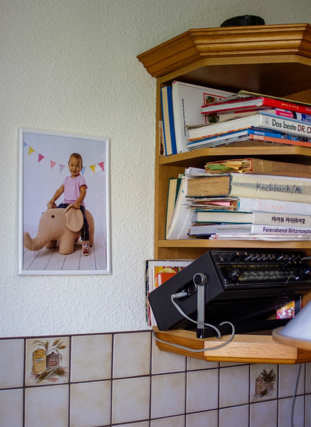Ein Regal mit Bücher, Notizzetteln und einem Radio, an der Wand hängt ein Foto eines lachenden Mädchens, das auf einem Kuschel-Elefanten sitzt.