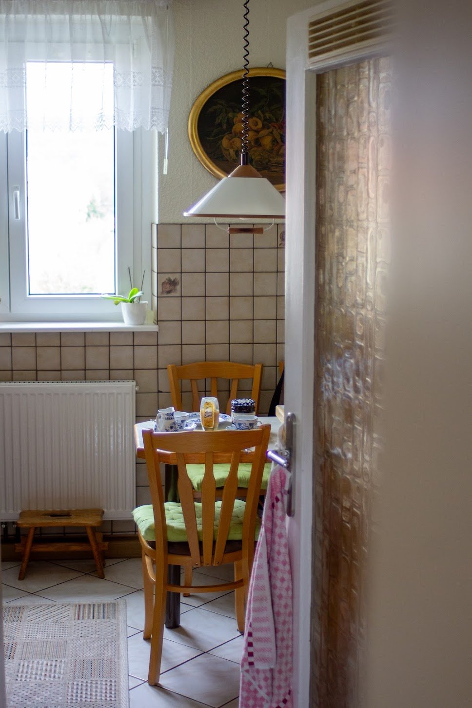 Kleine Küche mit Tisch und Stühlen, Vasen, Fenster mit Vorhängen und Pflanzen, Blick durch Tür in den Essbereich bei Tageslicht.