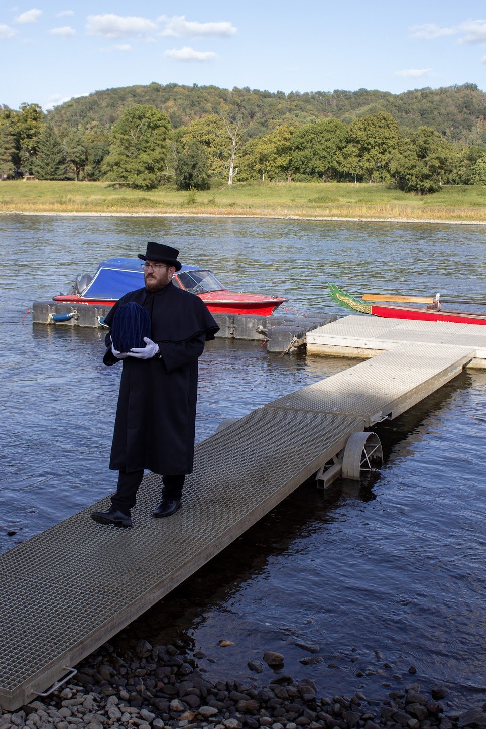 Ein Mann in dunkler Tracht mit Zylinderhut und Handschuhen trägt eine schwarze Kappe auf einem Steg an einem See, im Hintergrund sind Boote und ein Baum an einem bewaldeten Hügel bei sonnigem Himmel.