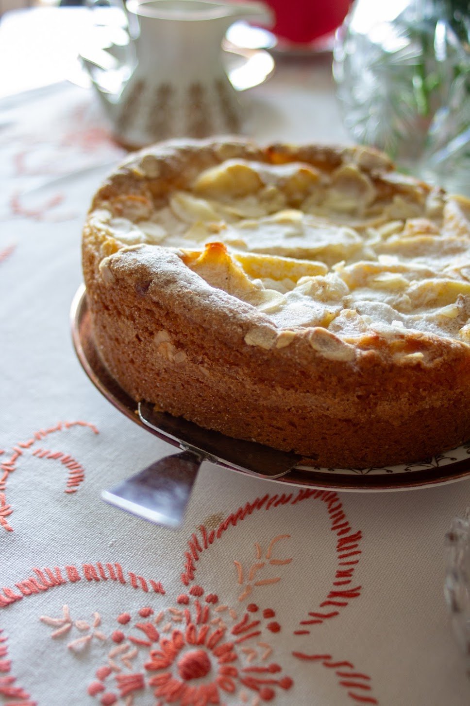 Ein Apfelkuchen auf einem weißen Tischtuch mit rotem Stickmuster. Im Hintergrund sind eine weiß getöpferte Teekanne und andere Gegenstände. Der Kuchen hat eine goldbraune Kruste und ist mit Puderzucker bestäubt.
