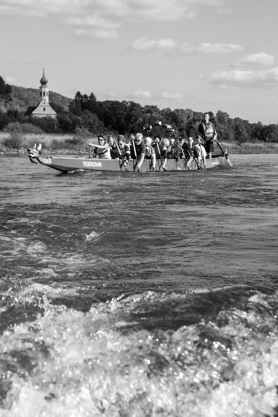Schwarz-Weiß-Foto einer Gruppe von Menschen, die in einem Boot namens "Tigerzahn" auf einem Fluss Rudern, mit einer Person am Steuer. Im Hintergrund sind Bäume und ein Gebäude mit einer Kirchturmspitze sichtbar.