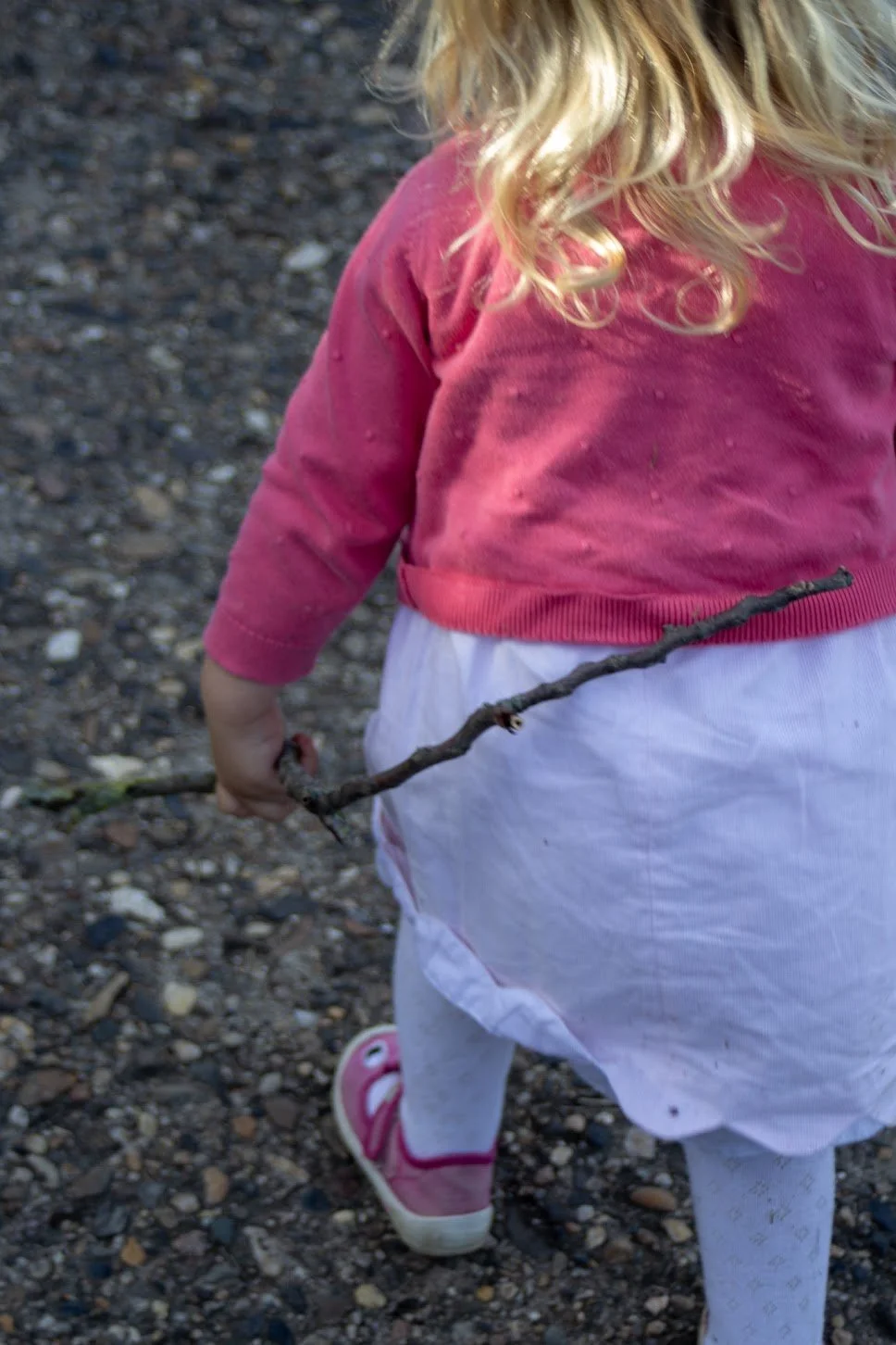 Ein Kind trägt eine pinke Jacke, weiße Rocke und pink-weiße Schuhe, hält einen dünnen Stock in der Hand, auf unbefestigtem Boden bestehend aus Kieselsteinen.