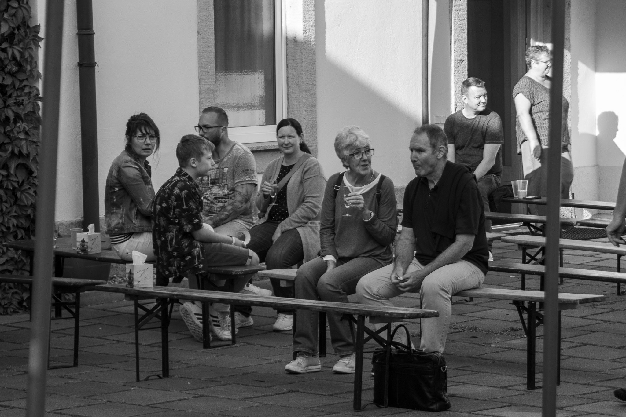 Mehrere Menschen sitzen auf Bänken im Außenbereich, unterhalten sich und trinken Getränke. Es sind Männer, Frauen und Kinder, die Atmosphäre wirkt entspannt und sozial.