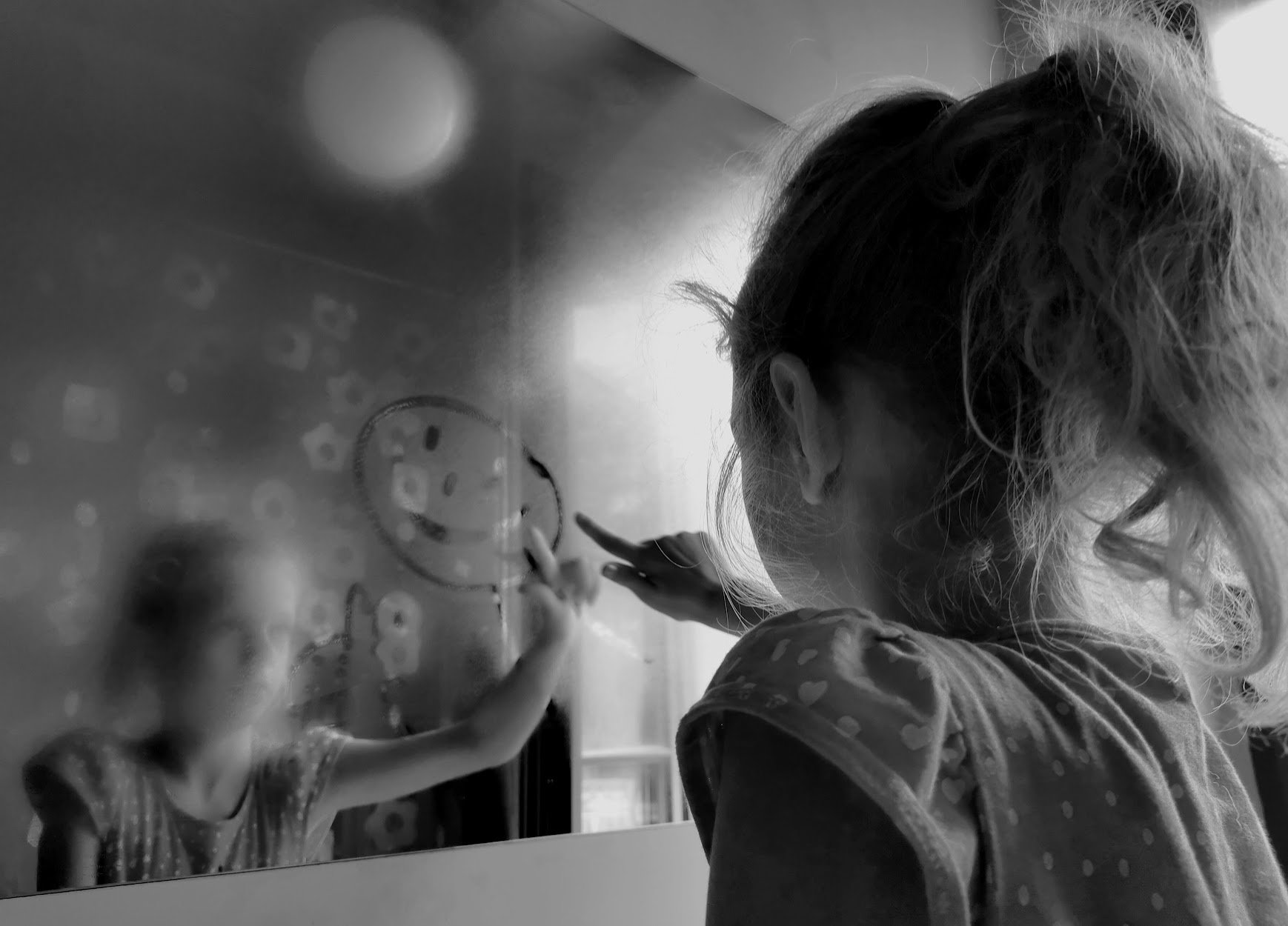 Ein Mädchen schreibt mit Finger auf einer Glasscheibe, im Hintergrund ein Spiegelbild.