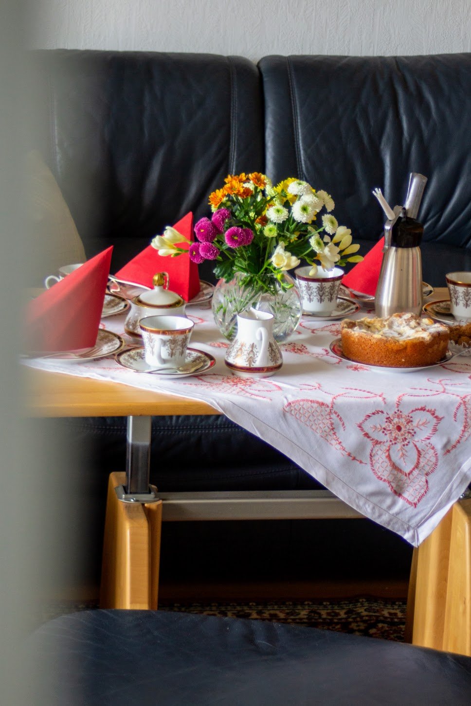 Ein gedeckter Frühstückstisch mit Blumenstrauß, Kaffee- und Teetassen, Kuchen und Servietten, auf schwarzem Ledersofa im Hintergrund.
