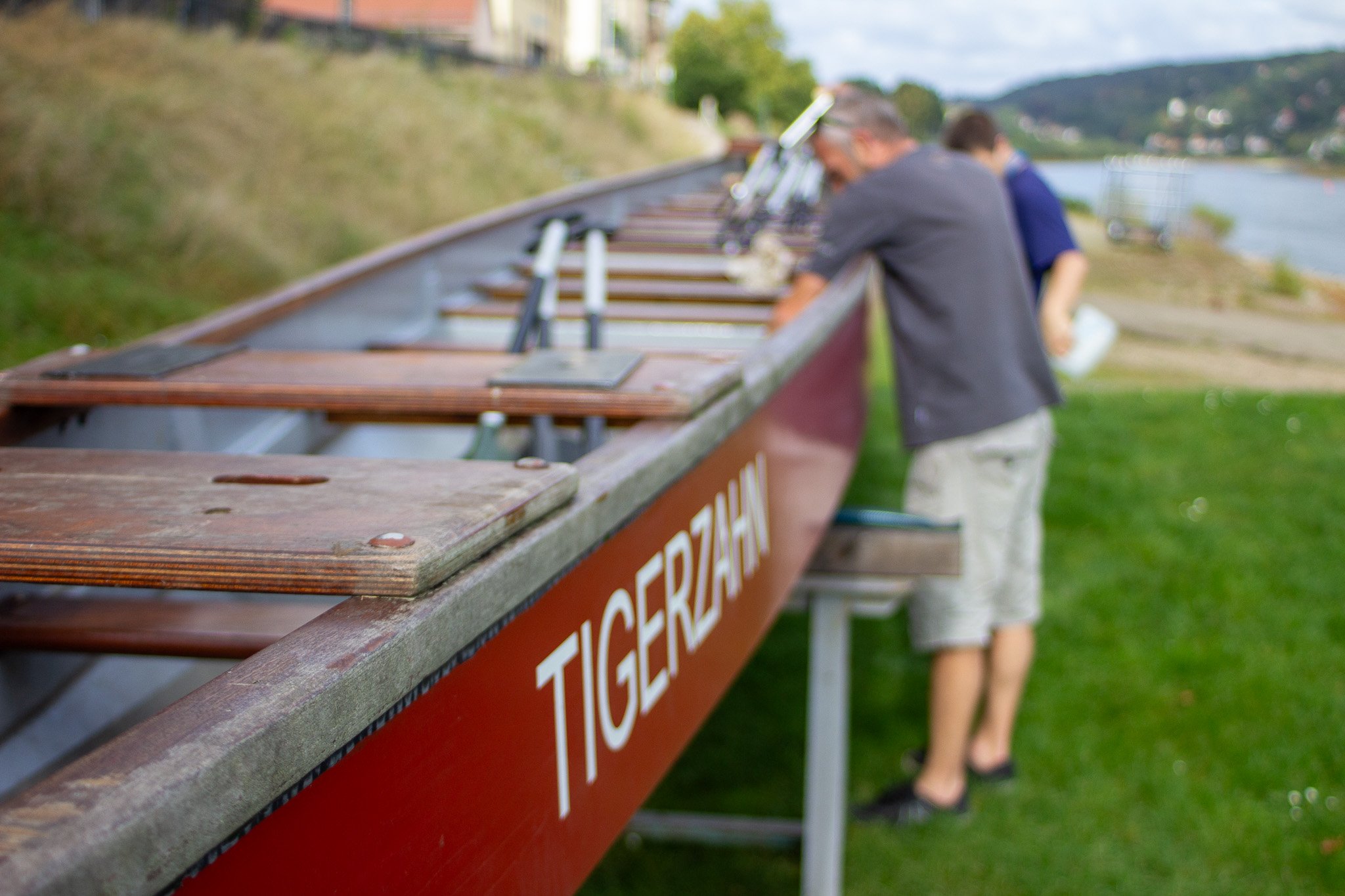 Blick auf ein Ruderboot mit der Aufschrift 'TIGERZAHN', im Hintergrund zwei Menschen, die sich auf das Boot vorbereiten, am Flussufer, bei schönem Wetter.